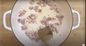 Cooking bacon cubes in a white pan, using a wooden spatula on a wooden cutting board background.